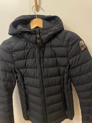 Svart dunjacka från Parajumpers - Säljer en svart, quiltad dunjacka från Parajumpers med huva och dragkedja framtill. Jackan har en patch på ärmen, innerficka märkt 'Security Pocket' och är superlätt. Perfekt för kyliga dagar och har en clean, sportig look. Knappast använd inga hål inga skador
