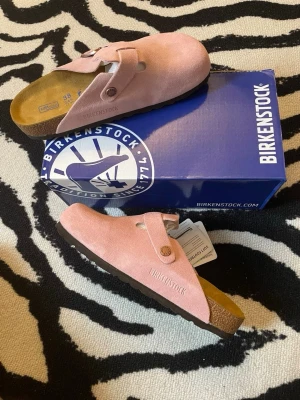Rosa mocka loafers från Birkenstock - Säljer ett par rosa loafers från Birkenstock i mjuk mocka med klassisk slip-in design och justerbart spänne i metall. Skorna har en bekväm fotbädd och robust sula, perfekta för dig som gillar både stil och komfort. Modellnamnet syns på sidan av skon.