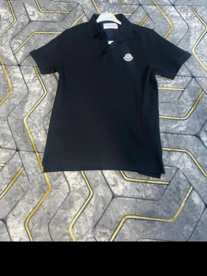 Svart Moncler pikétröja - Klassisk svartvit vitt  pikétröja från Moncler med liten logga på bröstet. Tröjan har krage, knappar framtill och korta ärmar. Perfekt till jeans eller shorts för en clean och stilren look.