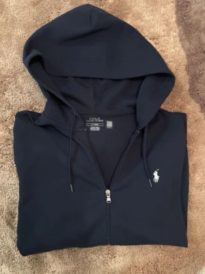 Polo Ralph Lauren - hoodie  - Snygg marinblå hoodie med dragkedja från Polo Ralph Lauren. Klassisk design med huva och justerbara snören, samt den ikoniska vita logon broderad på bröstet. Tillverkad i mjukt bomullsmaterial som är skönt att bära. Perfekt för en avslappnad och stilren look. Pris kan diskuteras vid snabbaffär