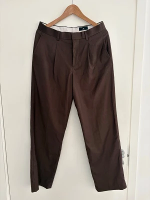 Bruna kostymbyxor från H&M - Snygga bruna kostymbyxor från H&M i regular fit. Byxorna har klassisk rak passform, pressveck och sidofickor. Perfekta för en stilren look och passar till skjorta eller t-shirt. Materialet känns mjukt och lätt, vilket ger en bekväm känsla hela dagen.