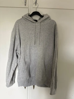 Ljusgrå basic hoodie  - En enkel och stilren ljusgrå hoodie med huva och dragsnören. Hoodien har en klassisk känguruficka framtill och ribbade muddar vid ärmslut och nederkant. Perfekt för en avslappnad och casual look. Bra skick 