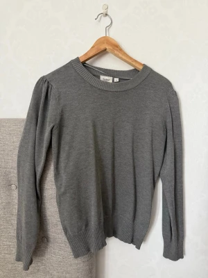 Grå stickad tröja från Saint - En enkel och stilren grå stickad tröja från Saint. Tröjan har rund halsringning, långa ärmar och ribbade muddar vid ärmslut och nederkant. Perfekt att matcha med jeans eller kjol för en clean look.