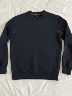 Louis vuitton Svart - Mörkblå sweatshirt från Louis Vuitton med broderad LV-logga på bröstet. Tröjan har rund halsringning, ribbade muddar och en snygg detalj med mönstrat tyg på insidan av kragen. Perfekt för en stilren och avslappnad look.