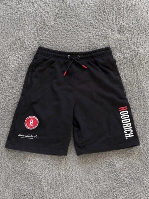 Svarta shorts från Hoodrich med tryck - Svarta shorts från Hoodrich med vit och röd logga på vänster ben och texttryck längs höger ben. Shortsen har snörning i midjan och sidofickor. Perfekta för en avslappnad streetwear-look.
