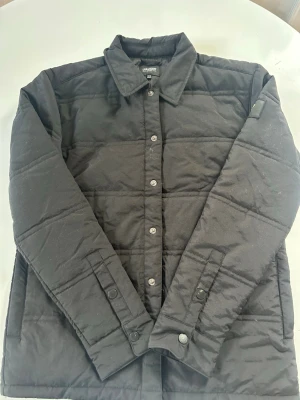 Svart quiltad overshirt - Stilren svart quiltad overshirt från Lyle & Scott med tryckknappar framtill och klassisk krage. Jackan har raka sömmar, diskret logga på ärmen och är lätt vadderad för extra värme. Perfekt lager-på-lager-plagg för kyligare dagar.