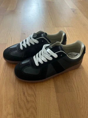 Svarta sneakers med vita detaljer - Snygga svarta sneakers med vita skosnören och beige insida. Skorna har en klassisk låg siluett och ovandel i mocka och läder. Yttersulan är i gummi och ger bra grepp. Perfekta för en stilren och avslappnad look.