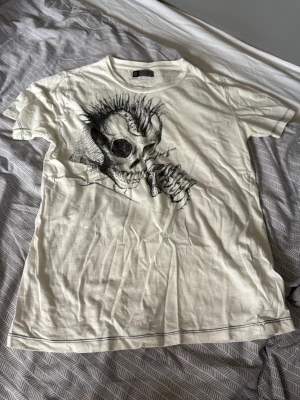 Vit t-shirt med dödskalletryck - Säljer en vit t-shirt med ett stort svart tryck av en dödskalle med mohawk på framsidan. T-shirten har korta ärmar och rund hals. Materialet känns som bomull och modellen är normal i passformen. Perfekt för dig som gillar edgy och alternativ stil.