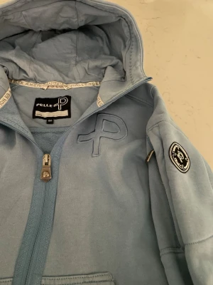 Ljusblå hoodie från Pelle P - Ljusblå hoodie från Pelle P med dragkedja framtill, stor huva och två fickor. Broderad P-logga på bröstet och ett runt märke på ärmen. Tillverkad i mjukt bomullsmaterial, perfekt för en avslappnad stil. Har ett litet märke på ärmen, knappt synligt.