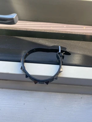 Valentino armband - Säljer ett svart armband i konstläder med metallnitar i pyramidform. Armbandet har justerbart spänne i metall och passar perfekt för en edgy eller alternativ stil. Enkel att matcha med andra accessoarer och ger en cool touch till din outfit. En tidig ägare och sägs har köpt i harrods, london