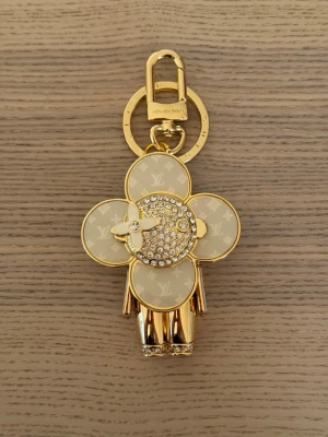 Vuitton guldfärgad nyckelring - Exklusiv nyckelring från Vuitton i guldfärgad metall med ikoniskt Monogram-mönster och gnistrande stenar. Designad som en blomfigur med vita och guldiga detaljer, perfekt som accessoar till väskan eller nycklarna.