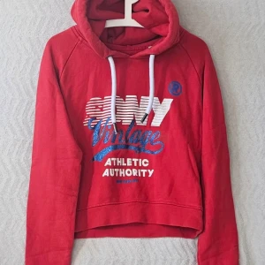 Röd hoodie LA By SDry med tryck - Snygg röd hoodie från LA By SDry med vit och blå text på bröstet där det står 'Vintage Athletic Authority'. Hoodien har huva med vita snören och en avslappnad passform. Perfekt för en sportig och casual stil.