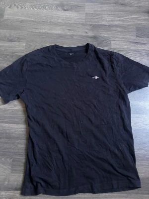 Svart t-shirt från GANT - Klassisk svart t-shirt från GANT med rund halsringning och liten broderad logga på bröstet. Tillverkad i mjuk bomull som känns skön mot huden. Perfekt basplagg för alla tillfällen och enkel att matcha med andra plagg.