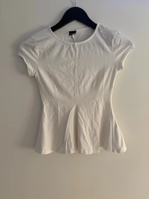 Vit figursydd topp med kort ärm - Snygg vit topp med rund halsringning och korta ärmar. Modellen är figursydd med lätt utsvängd nederdel som ger en feminin siluett. Perfekt att styla med jeans eller kjol för en fräsch look.
