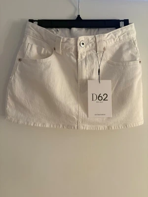 Vit kort jeanskjol med fickor - Snygg vit kort jeanskjol med klassiska fem fickor, bälteshällor och knappstängning fram. Kjolen har en rak passform och är perfekt för en trendig och avslappnad stil. Passar till allt från sneakers till boots.