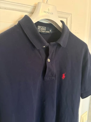 Mörkblå pikétröja från Ralph Lauren - Klassisk mörkblå pikétröja från Polo Ralph Lauren med röd häst på bröstet. Tröjan har krage och två knappar framtill. Tillverkad i mjukt bomullsmaterial för en bekväm känsla.