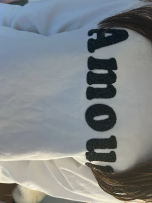 Vit hoodie med svart text Amour - Säljer en vit hoodie med stor svart text 'Amour' i glittrande material på ryggen. Hoodien har en klassisk huva och är gjord i mjukt tyg, perfekt för en avslappnad och trendig look.