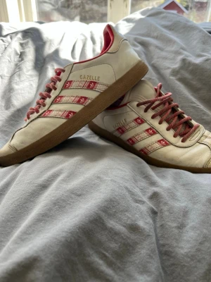 Adidas Gazelle vita och röda sneakers - Säljer ett par Adidas Gazelle sneakers i vitt läder med röda detaljer och mönstrade ränder. Skorna har röda snören och en klassisk gummisula i brunt. Perfekta för dig som gillar retrostil och vill sticka ut med snygga färgkombos. Finns en liten skada på sulan vid hälen men det är inget hål bara att det har släppt lite. Skriv vid intresse