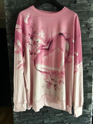 Rosa sweatshirt med hjärtmotiv - En oversized rosa sweatshirt med stort hjärtmotiv och blommiga detaljer framtill. Tröjan har långa ärmar, rund halsringning och mjukt material. Perfekt för dig som gillar en söt och romantisk stil.