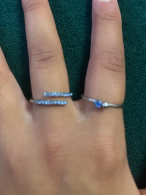 Två silvriga ringar med blå stenar - Säljer två stilrena silvriga ringar, en med en enkel blå sten och en öppen ring med flera små blå stenar på rad.