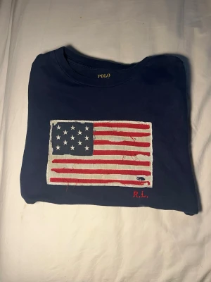 Blå t-shirt Polo Ralph Lauren USA-flagga - Mörkblå t-shirt från Polo Ralph Lauren med en stor broderad amerikansk flagga på bröstet och RL-signatur i rött. Klassisk rund hals och normal passform. Tillverkad i mjuk bomull som känns skön mot huden.