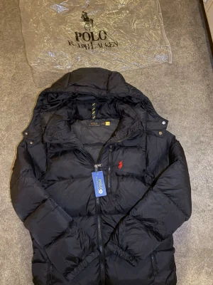 Dunjacka Polo Ralph Lauren Svart med Röd Pony 🐎 - Helt ny Polo Ralph Lauren dunjacka i klassisk svart färg med den ikoniska röda ponnylogotypen broderad på bröstet. Jackan är en varm och stilren pufferjacka, perfekt för kallare väder, och kommer med alla originaltaggar kvar.  ⭐️ Condition : 10/10 (New with tags) 🎲 Size : S 🎨 Color : Black 📦 Fast and careful shipping