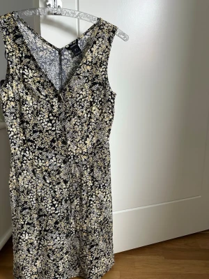 Blommig ärmlös kortklänning - Säljer en söt ärmlös kortklänning med svart botten och små vita och beige blommor. Klänningen har v-ringning både fram och bak och är tillverkad i ett lätt tyg som passar perfekt för varmare dagar. Figurnära passform och klassisk siluett. Aldrig använd dock tvättad en gång 