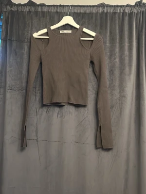 Brun ribbad långärmad topp - Trendig brun ribbad topp med långa ärmar och coola cut-out-detaljer vid axlarna. Toppen har en croppad passform och slits vid ärmsluten, vilket ger en modern vibe. Perfekt att styla med jeans eller kjol för en snygg look.