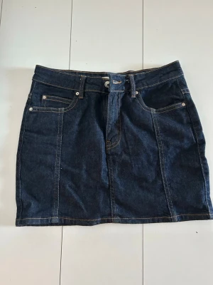 Mörkblå jeanskjol med fickor - Snygg mörkblå jeanskjol i klassisk modell med fem fickor, bälteshällor och kontrastsömmar. Kjolen har dragkedja och knapp framtill samt två stora bakfickor. Perfekt basplagg i garderoben för en avslappnad look.