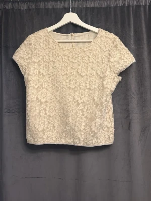 Beige spetsblus med pärlknappar - Söt beige blus med korta ärmar och vackert blommigt spetsmönster. Toppen har rund halsringning och pärlknappar längs ryggen som ger en elegant touch. Perfekt för dig som gillar romantiska detaljer och vill sticka ut lite extra.