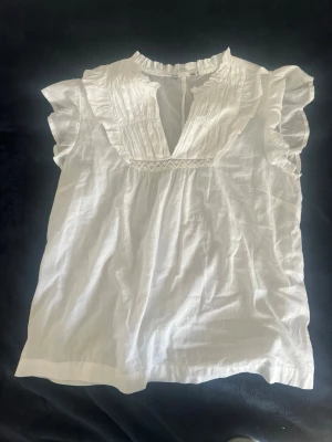 Vit blus med volang och v-ringning - Söt vit blus i luftig bomull med volangdetaljer runt halsen och v-ringning. Blusen har korta ärmar med lätt rynk och ett romantiskt uttryck. Perfekt till jeans eller kjol för en fräsch look.