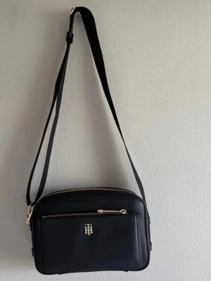 Svart axelväska Tommy Hilfiger - Ny axelremsväska från Tommy Hilfiger, endast använd någon enstaka gång. Justerbar axelrem, guldfärgade dragkedjor och TH-logga framtill. 