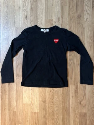 Svart långärmad topp Comme des Garçons - Svart långärmad topp från Comme des Garçons PLAY med det ikoniska röda hjärta-märket broderat på bröstet. Klassisk rund halsringning och mjukt bomullsmaterial som känns skönt mot huden. Perfekt för en clean och stilren look.