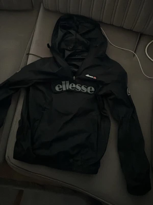 Svart vindjacka från Ellesse - Svart vindjacka från Ellesse med stor logga framtill och huva. Jackan har dragkedja upptill och är tillverkad i ett lätt, vindtätt material. Perfekt för blåsiga dagar och snygg streetstil. Klassisk passform med elastiska muddar.
