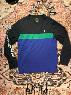 Långärmad topp från Polo Ralph Lauren - Snygg långärmad topp från Polo Ralph Lauren med svart överdel, grön rand och blå nederdel. Stor logga på ena ärmen och liten broderad logga på bröstet. Perfekt för dig som gillar sportig och avslappnad stil.
