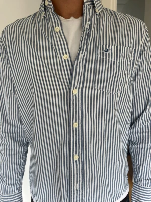 Blåvit randig skjorta med ficka - Säljer en klassisk blåvit randig skjorta med knappar framtill och bröstficka. Skjortan har button down-krage och långa ärmar. Tillverkad i bomull, perfekt för en avslappnad men stilren look. Model 185 cm, 71kg. Hör av er vid frågor