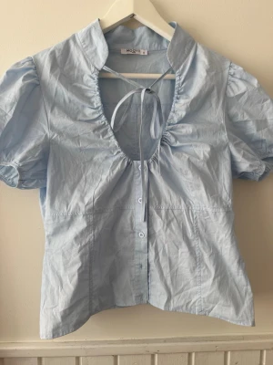 Ljusblå blus med puffärm och knyt - Supersöt ljusblå blus från MOVA med puffärmar, knappar framtill och snygg knytning vid halsen. Djup urringning och lätt figursydd passform. Perfekt till jeans eller kjol för en trendig look.
