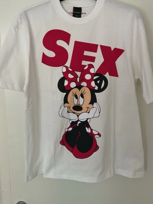 Vit t-shirt med Minnie Mouse-tryck - Unik vit t-shirt från FAR ARCHIVE i storlek M med stort tryck av Minnie Mouse och texten 'SEX' i rött. T-shirten har korta ärmar och rund hals. Materialet är mjuk bomull och trycket är i rött, svart och vitt med detaljer i beige.