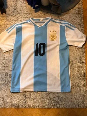 Argentina fotbollströja Messi #10 - Säljer en klassisk Argentina landslagströja med ljusblå och vita ränder, nummer 10 och Messi på ryggen. Tröjan har korta ärmar, V-ringning och tre stjärnor ovanför landslagsemblemet. Perfekt för dig som älskar fotboll och Messi.