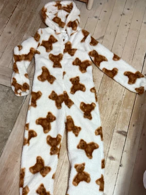 Onesie med teddybjörnar och huva - Supermjuk och fluffig onesie i vitt med brunt teddybjörnsmönster. Heltäckande dragkedja framtill och huva. Perfekt för mysiga hemmakvällar och extra värme. Långa ärmar och ben för maximal komfort.