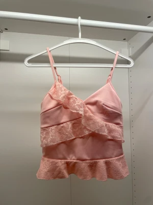 Rosa pyjamastopp med spetsdetaljer - Söt pyjamastopp i ljusrosa satin med tunna axelband och volangkant. Toppen har dekorativa spetsdetaljer framtill och längs nederkanten som ger en romantisk känsla. Perfekt för mysiga kvällar hemma.
