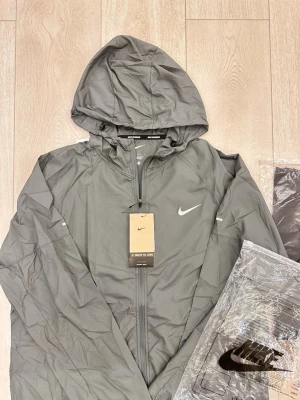 Grå vindjacka med huva från Nike - Snygg grå vindjacka från Nike med dragkedja framtill och justerbar huva. Jackan har en minimalistisk design med Nike-logga på bröstet och är tillverkad i lätt, vindtätt material. Perfekt för blåsiga dagar och enkel att matcha med olika outfits.