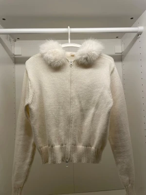 Vit stickad hoodie från Gina Tricot - Supersöt vit stickad hoodie från Gina Tricot med dragkedja framtill och fluffiga fuskpälsbollar på huvan. Tröjan har ribbade muddar och är perfekt för dig som vill ha en mysig och trendig look. Materialet känns mjukt och varmt.