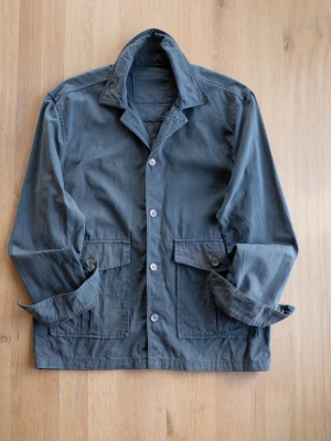 Green/Blue Chore Jacket, size M - Stilren grön/blå utility jacka i bomull med klassisk krage och knäppning framtill. Två stora fickor med lock och knapp framtill ger en cool och funktionell look. Perfekt för lager på lager och enkel att matcha till olika outfits.