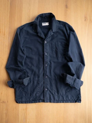 Navy Chore Jacket, size M - Snygg mörkblå overshirt med klassisk krage och tre fickor framtill. Skjortan har långa ärmar och stängs med knappar. Perfekt att bära öppen eller stängd för en avslappnad look.