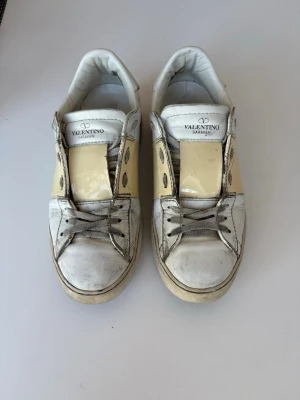 Valentino Garavani vita sneakers - Snygga vita sneakers från Valentino Garavani med beige detaljer och vita nitar på sidorna. Skorna har en klassisk låg siluett, rund tå och snörning framtill. Tillverkade i läder med en chunky vit sula för en trendig look. Pris kan diskuteras! 