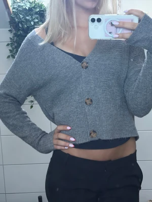 Grå cropped kofta med knappar - Säljer en grå, ribbstickad cropped kofta med stora bruna knappar framtill. Koftan har lång ärm och en avslappnad passform som passar perfekt till både jeans och kjol. Materialet känns mjukt och bekvämt mot huden.