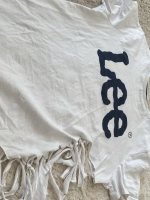 Vit waist t shirt lee. - Vit t-shirt i bomull med svart tryck framtill och knytfransar längs nederkanten. T-shirten har korta ärmar och en avslappnad passform. Perfekt för dig som gillar y2k , unik stil.