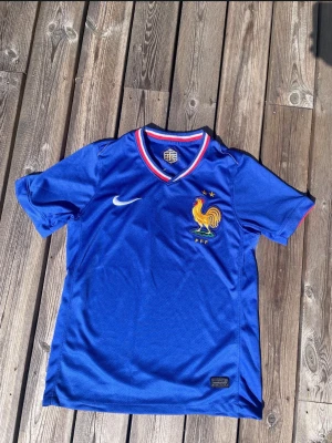 Blå Frankrike fotbollströja Nike - Säljer en blå fotbollströja från Nike med Frankrikes landslagsmärke och två stjärnor på bröstet. Tröjan har korta ärmar, rundad krage med röd och vit kant samt meshmaterial för bra ventilation. Perfekt för match eller träning.