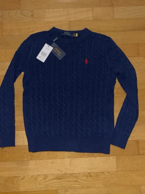 Marinblå kabelstickad tröja Polo Ralph Lauren - Kabelstickad marinblå tröja från Polo Ralph Lauren med rund halsringning och röd broderad logga på bröstet. Tröjan har långa ärmar och ribbade muddar vid ärmslut och nederkant. Perfekt för en stilren och klassisk look.